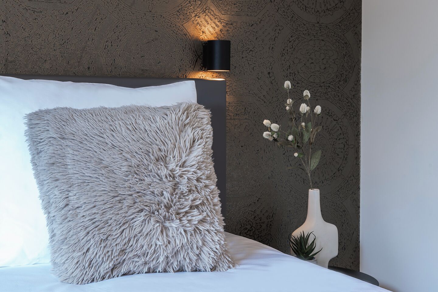 A bed with a fluffy pillow, a white vase with flowers, and a wall-mounted light fixture.