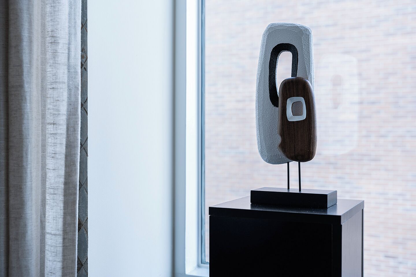 Une sculpture moderne, posée sur un piédestal près d’une fenêtre, avec un mur de briques en arrière-plan.