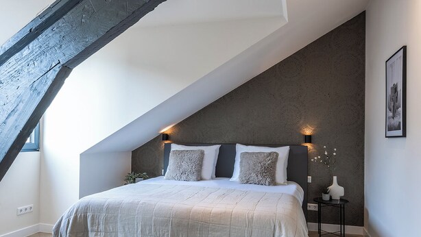 Ein Schlafzimmer mit schrägverdeckter Decke, einem Bett mit grauem Kopfteil und einem kleinen Nachttisch mit Vase und Pflanze.