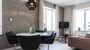 Ein modernes Wohnzimmer mit einem runden Esstisch, schwarzen Stühlen, einem grauen Sofa, einem Fernseher und einem großen Fenster mit Vorhängen.