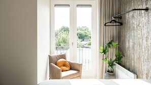 Ein Zimmer mit einem Bett, einem Sessel mit Kissen, einer Zimmerpflanze und Blick auf Grünfläche.