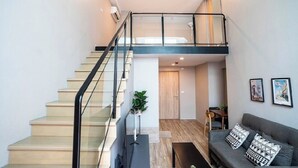 A modern apartment with a staircase leading to a loft bedroom, a living area with a sofa and cushions, and a dining area with a table and chairs.