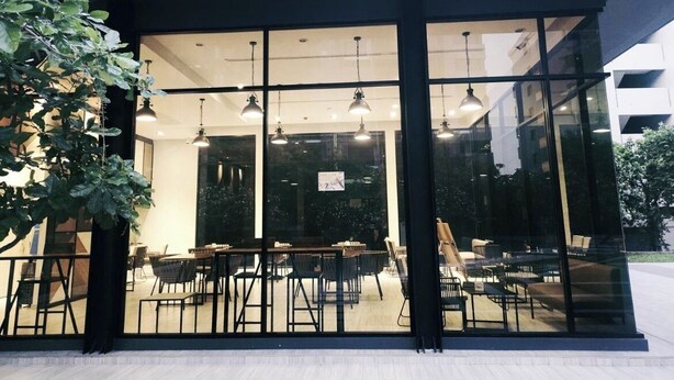 A modern building with large glass windows, outdoor seating area with tables and chairs, and hanging pendant lights.