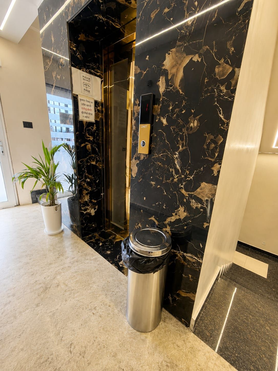 An elevator with a black marble wall, a trash bin, and a potted plant in a hallway.