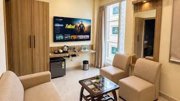 Uma sala de estar moderna com uma TV de tela plana exibindo um filme, uma mesa de centro, dois sofás e uma pequena área de cozinha.