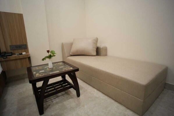 A small, neatly arranged room with a beige sofa, a glass-top table with a vase of flowers, and a wooden side table with a phone and charger. - Hotel Om Villa (Varanasi)