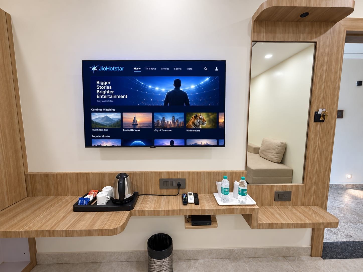 Zona de recepción de un hotel con un televisor de pantalla plana grande mostrando la interfaz de un canal de cine, un mostrador de madera con una cafetera, un cubo de basura y un espejo.