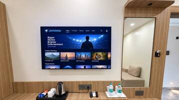 Zona de recepción de un hotel con un televisor de pantalla plana grande mostrando la interfaz de un canal de cine, un mostrador de madera con una cafetera, un cubo de basura y un espejo.