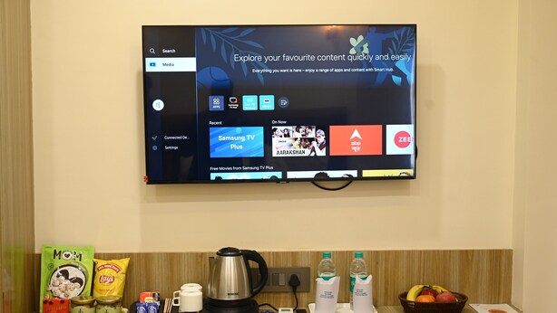 A hotel room with a TV displaying a media app interface, a coffee maker, snacks, and a fruit basket on the counter. - Hotel Om Villa (Varanasi)