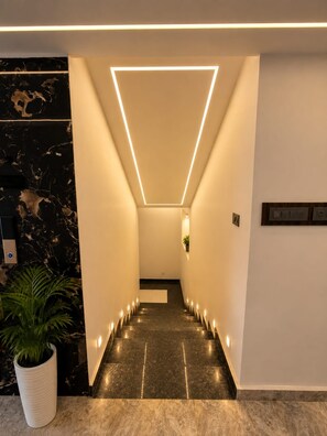 A modern hallway with a geometric light fixture, marble walls, and a potted plant. - Hotel Om Villa (Varanasi)