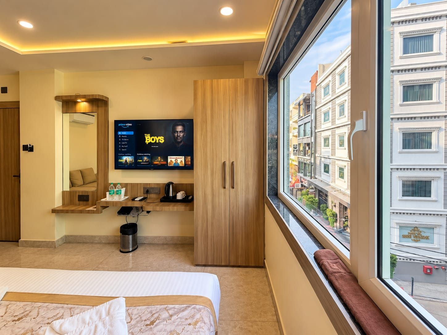Habitación de hotel con cama, televisor montado en la pared, ventana con vista a edificios y una pequeña mesa con hervidor y tazas.