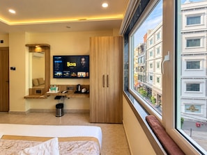 Habitación de hotel con cama, televisor montado en la pared, ventana con vista a edificios y una pequeña mesa con hervidor y tazas.