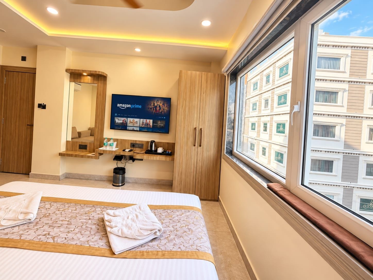 A hotel room with a large bed, a television mounted on the wall, a window with a view of buildings, and a wooden cabinet.