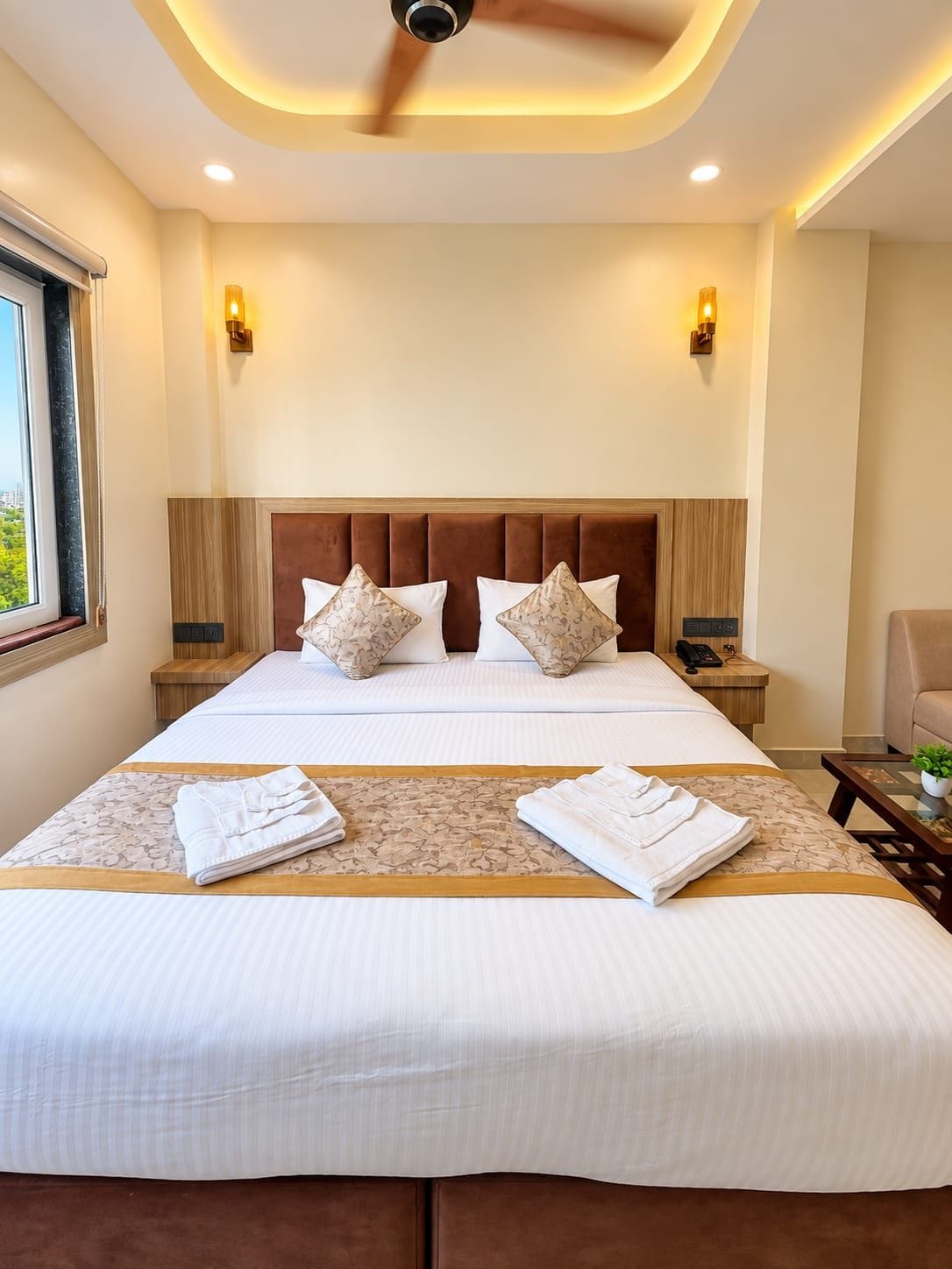 A hotel room with a large bed, two bedside tables, a ceiling fan, and a window with a view.