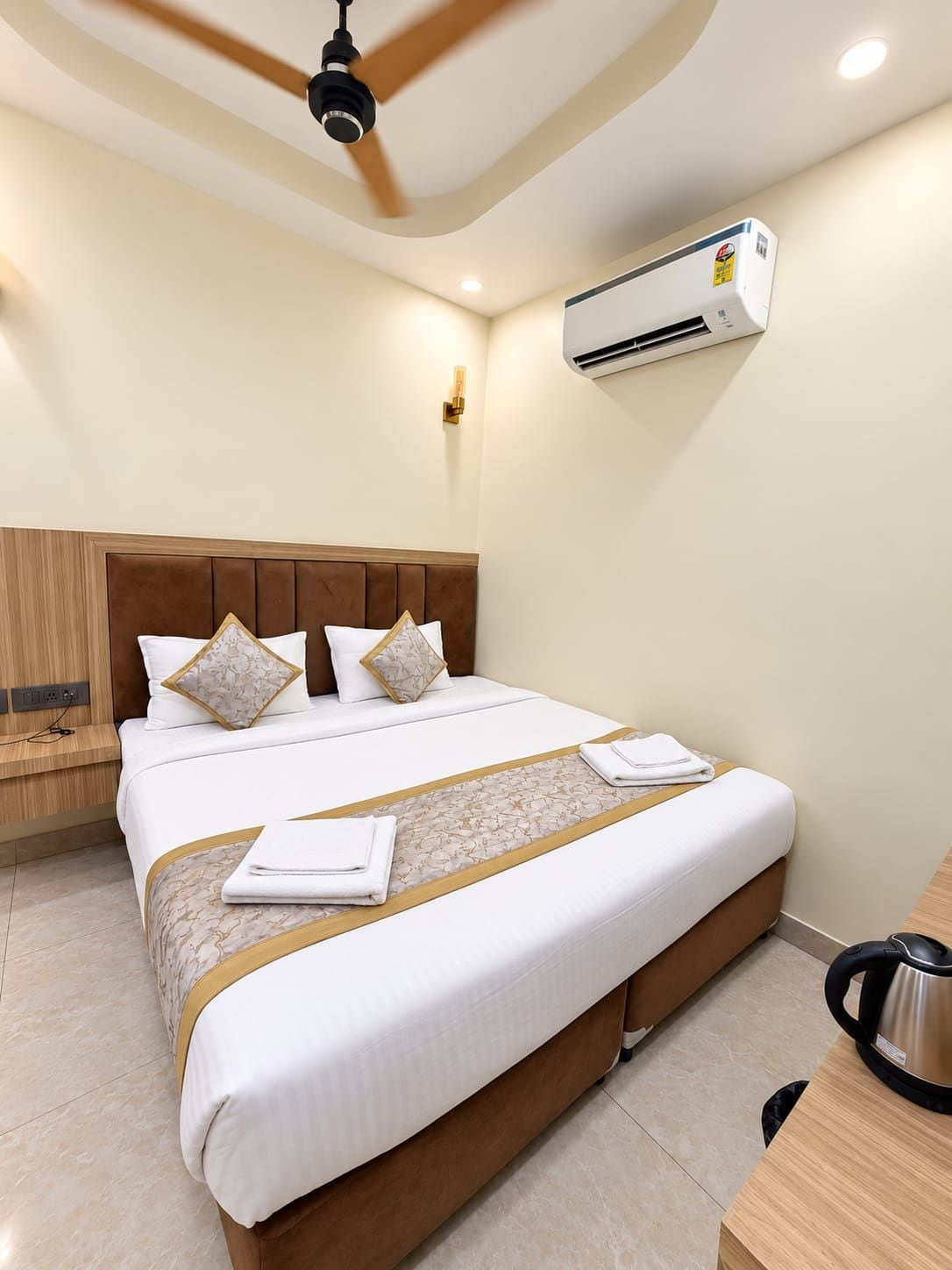Habitación de hotel con cama, ventilador de techo, equipo de aire acondicionado, mesita de noche y cafetera.