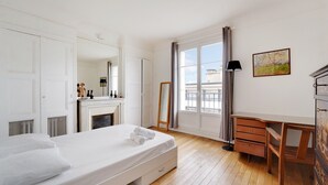 A bedroom with a bed, a dresser, a desk, a mirror, a fireplace, and a window with a view.