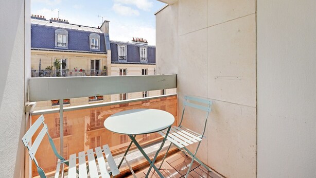A balcony with a metal chair and a small table, overlooking a row of buildings with blue facades and white windows.