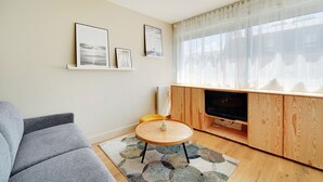 A living room with a grey sofa, a round wooden coffee table, a TV on a wooden cabinet, and a large window with sheer curtains.