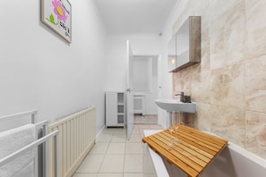 Un couloir étroit avec un radiateur blanc, un banc en bois, un lavabo blanc et une porte donnant sur une autre pièce.