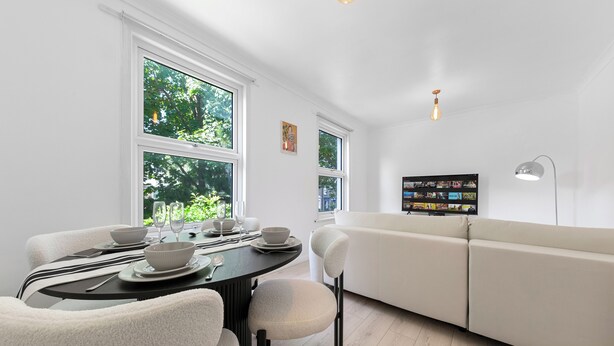 A modern dining area with a round table set for two, a white sofa, and a wall-mounted TV. - Charming London Apartment Near City Center (London)