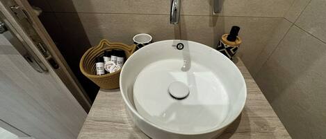 Un baño con un lavamanos de cerámica blanca, un mueble de tocador de madera y una cesta de mimbre con artículos de aseo.