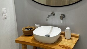 A bathroom with a white sink, two faucets, a wooden vanity, a mirror, a small basket with toiletries, and a cup. - Georgias House Project West (Skiathos)