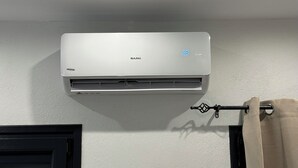 A white air conditioning unit mounted on a wall with a control panel and a remote control hanging on a nearby curtain rod. - Georgias House Project West (Skiathos)