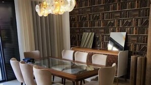 Een eetgedeelte met een tafel met glazen blad, omringd door stoelen, een kroonluchter en een boekenkast op de achtergrond.