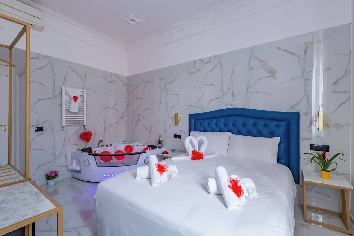 A hotel room with a marble-patterned wall, a blue headboard, a bed with white linens and red bows, a small table with a plant, and a massage chair with red accents.