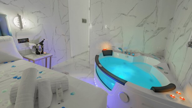 A modern bathroom with a marble wall, a bathtub with blue lighting, a small table with a bottle and glasses, and towels on a bed. - MISS SUITE  (Rome)