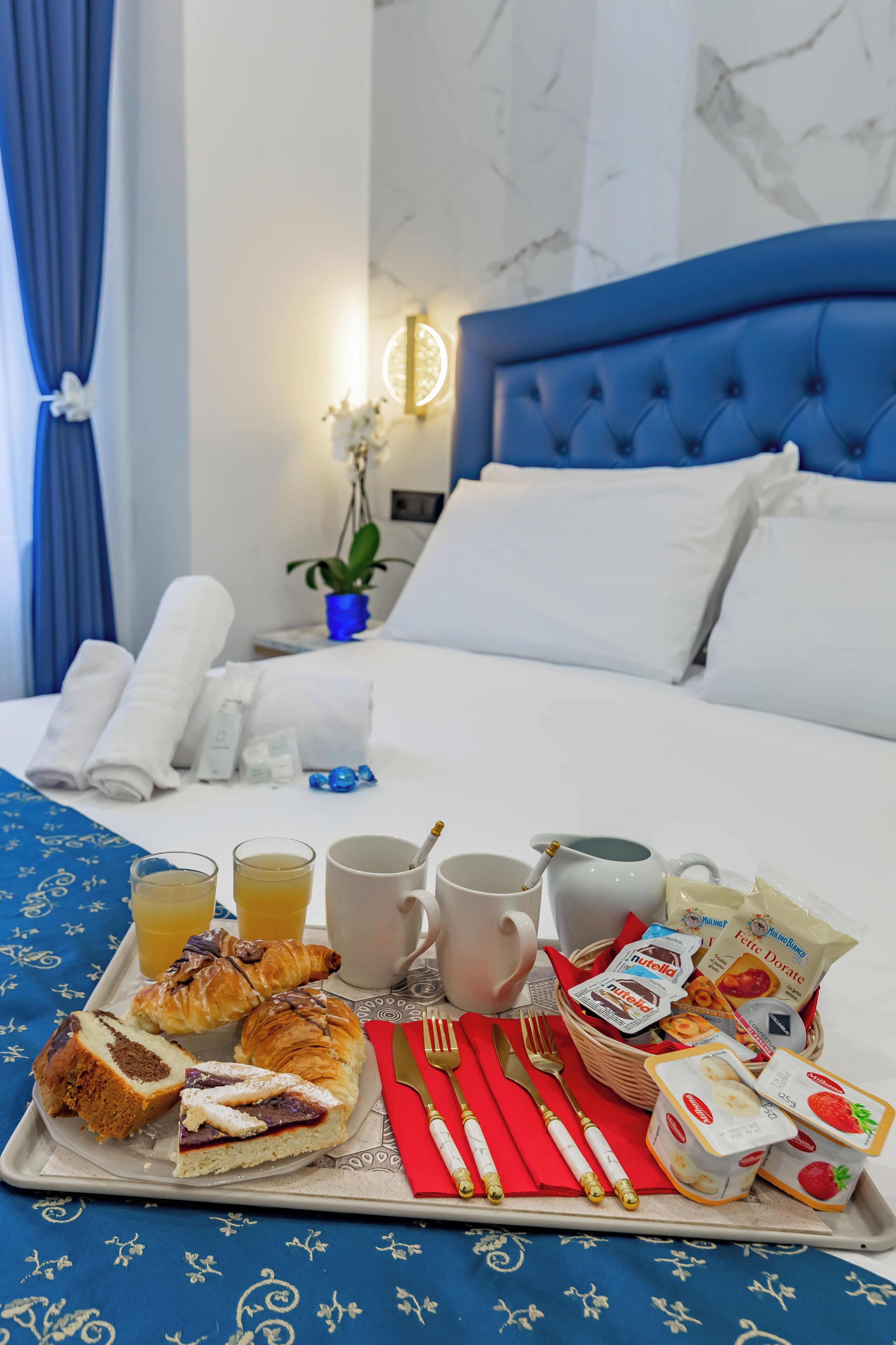 A neatly made breakfast tray with croissants, a slice of cake, and a bottle of juice on a bed with a blue headboard and white pillows.