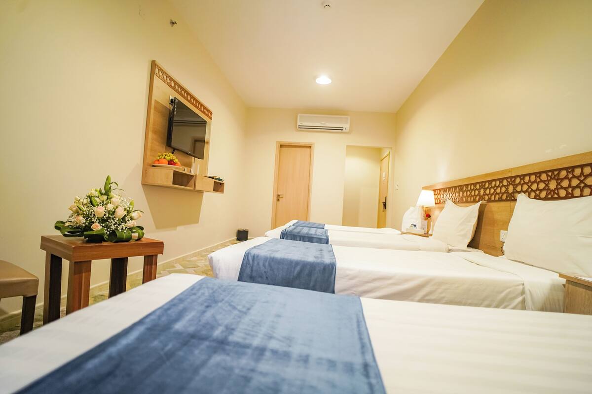Une chambre d’hôtel avec trois lits, une tête de lit en bois, un téléviseur fixé au mur, un appareil de climatisation et une petite table agrémentée d’un arrangement floral.