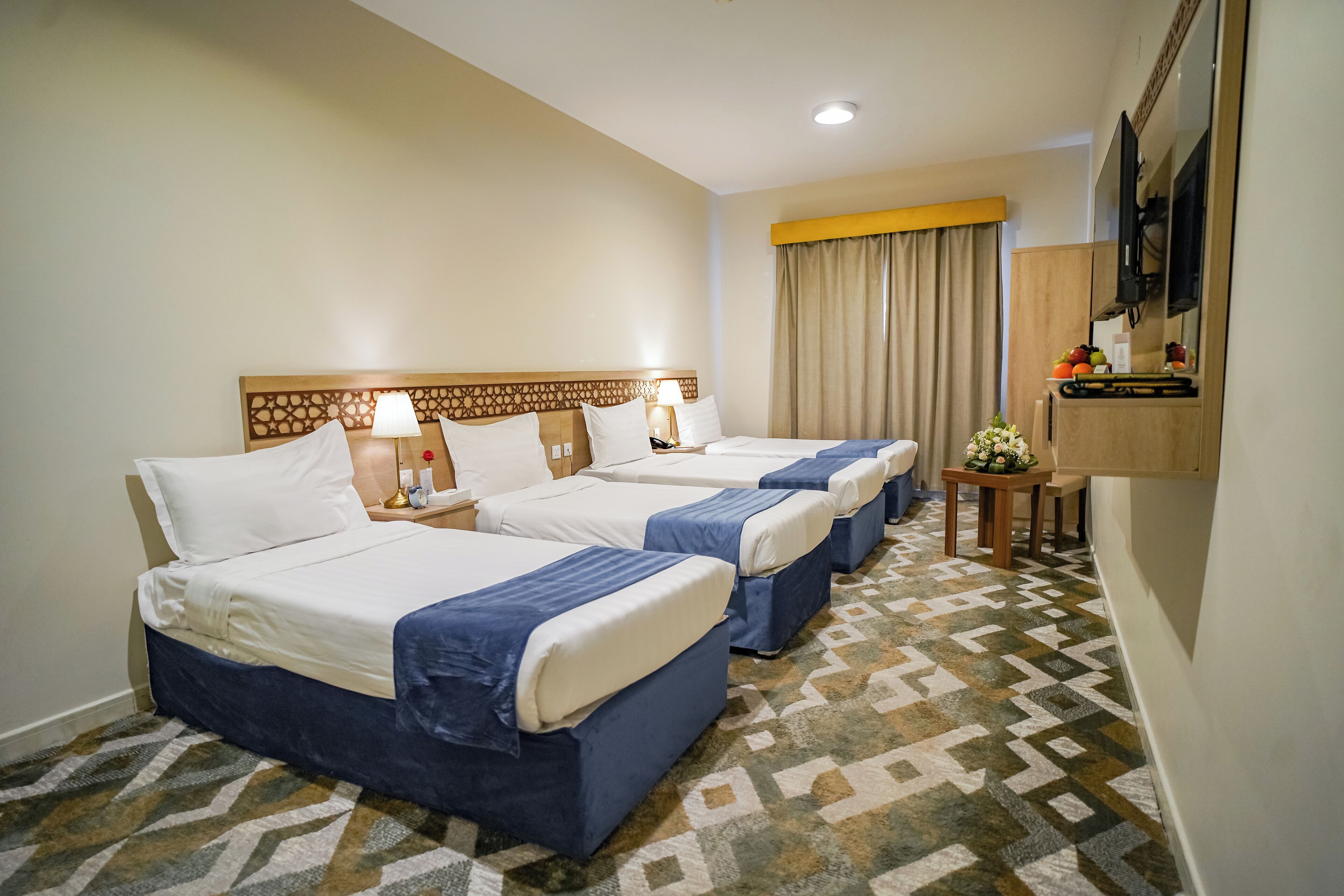 A hotel room with three beds, a small table with a vase of flowers, and a television mounted on the wall.