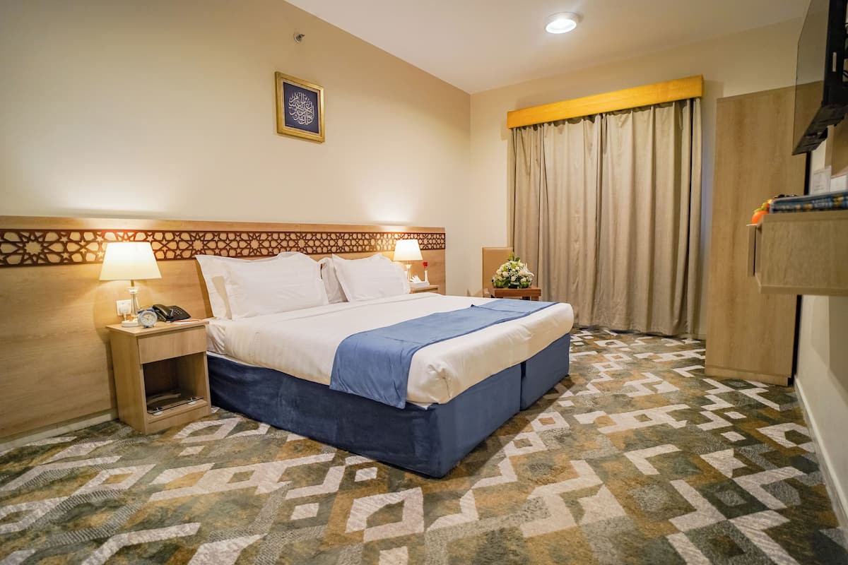 Une chambre d’hôtel avec un grand lit, deux tables de chevet avec des lampes, une tête de lit décorative, une moquette à motifs et une fenêtre avec des rideaux.