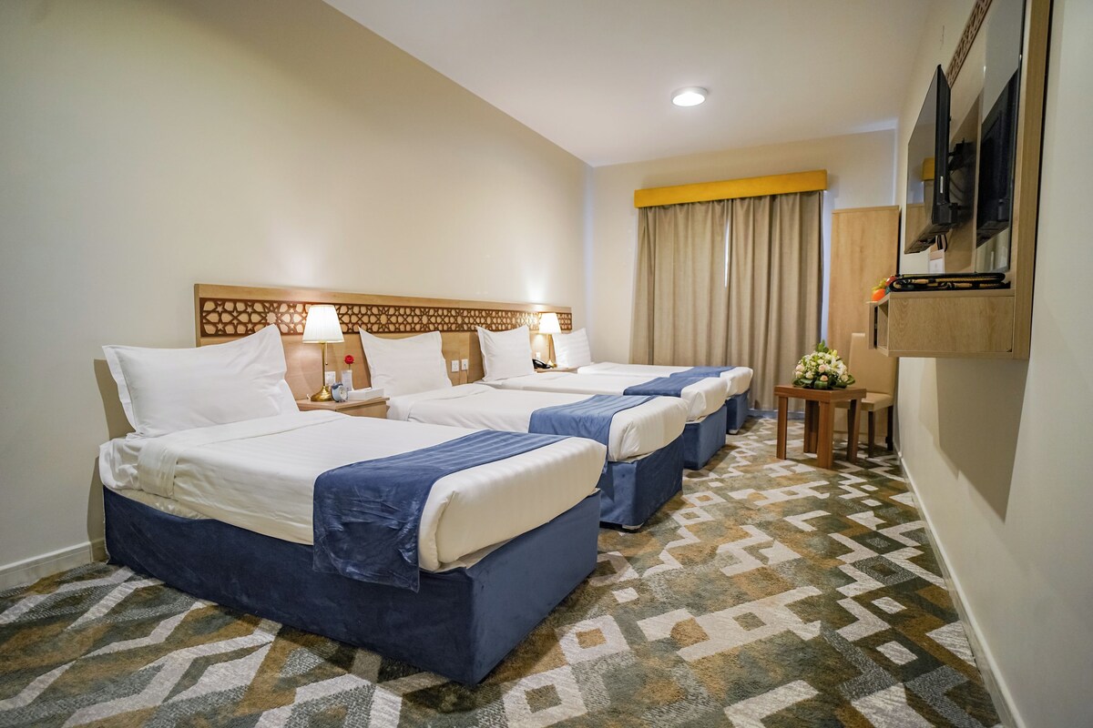 A hotel room with three beds, a TV, a small table with a vase, and a patterned carpet.