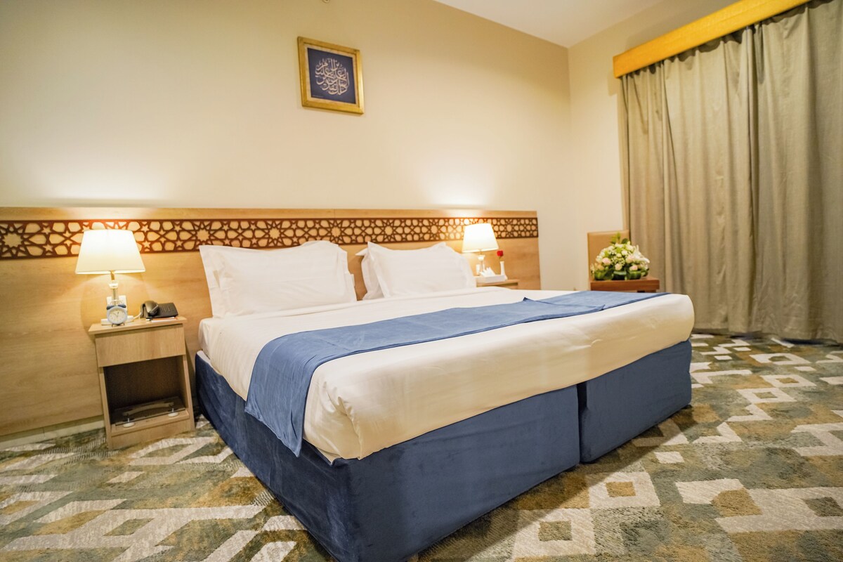 A hotel room with a large bed, two bedside tables with lamps, a decorative headboard, a framed artwork on the wall, and a patterned carpet.