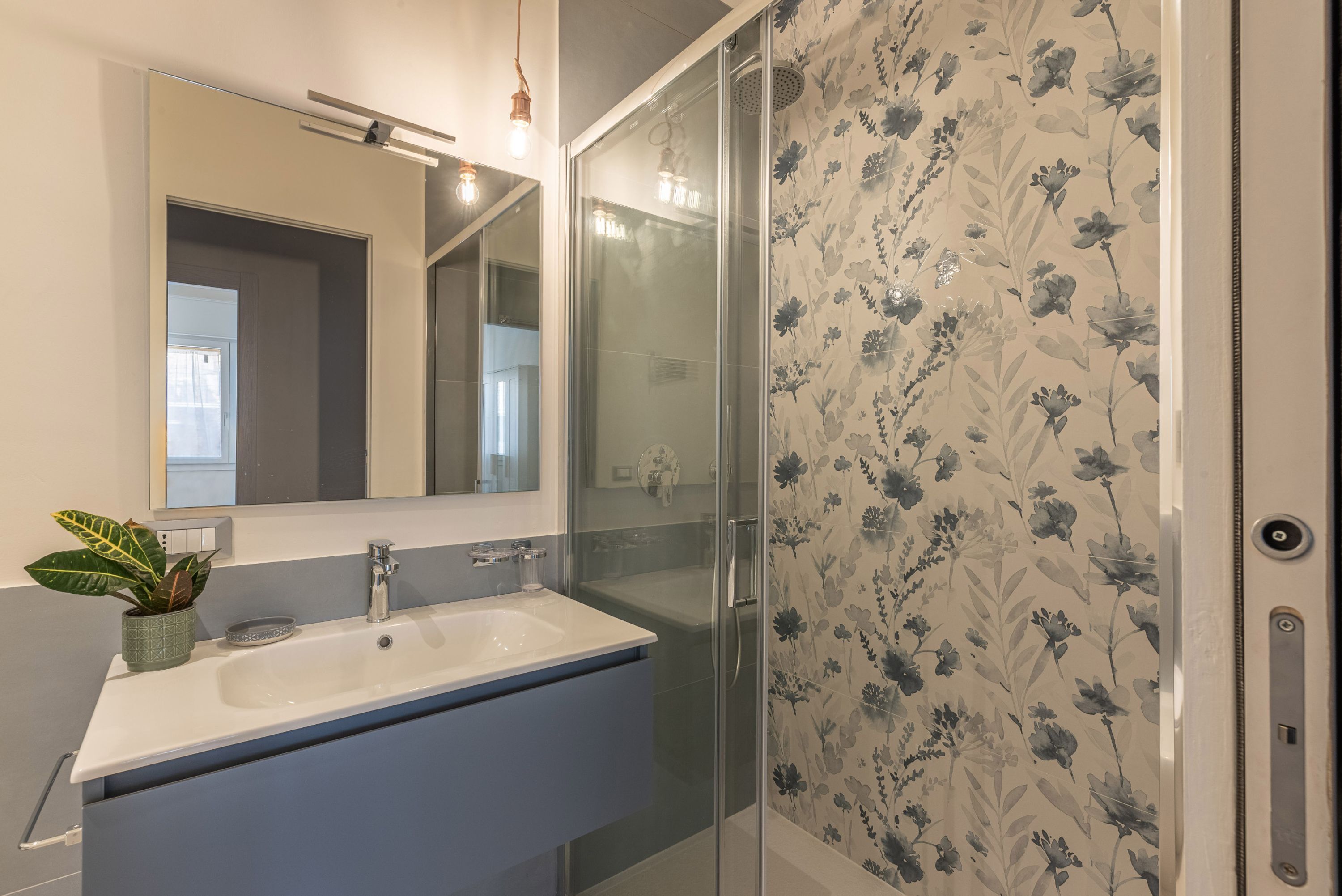 A bathroom with a white sink, a mirror, a shower with glass doors, and a floral wallpaper.
