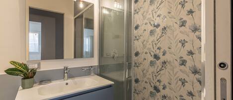 A bathroom with a white sink, a mirror, a shower with glass doors, and a floral wallpaper.