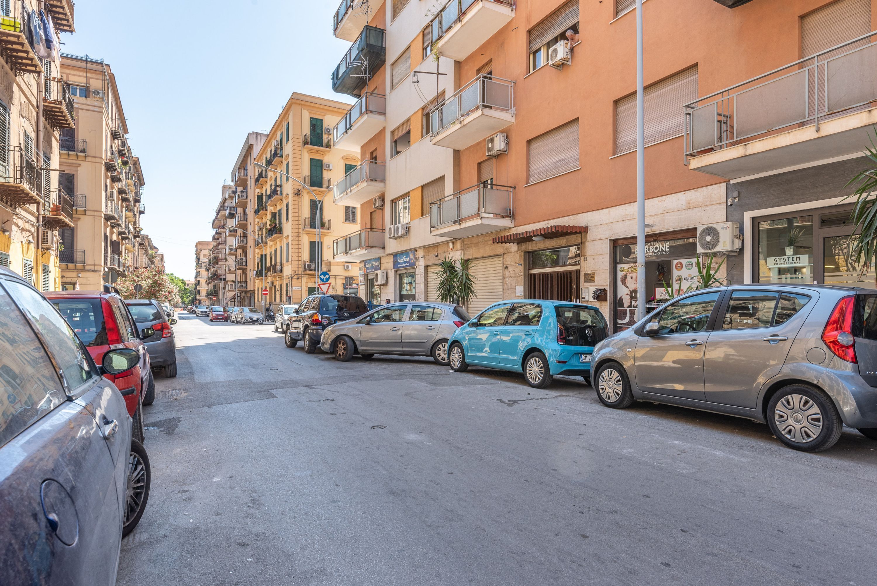 A street lined with multi-story residential buildings, parked cars, and a clear sky.