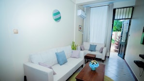A living room with a white sofa, a wooden coffee table, and a small side table. There is a window with curtains and an air conditioning unit on the wall. - Kilimanjaro Home Of Travelers (Moshi)