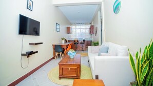 A modern living room with a flat-screen TV, a dining area with chairs, a sofa, and a kitchen in the background. - Kilimanjaro Home Of Travelers (Moshi)