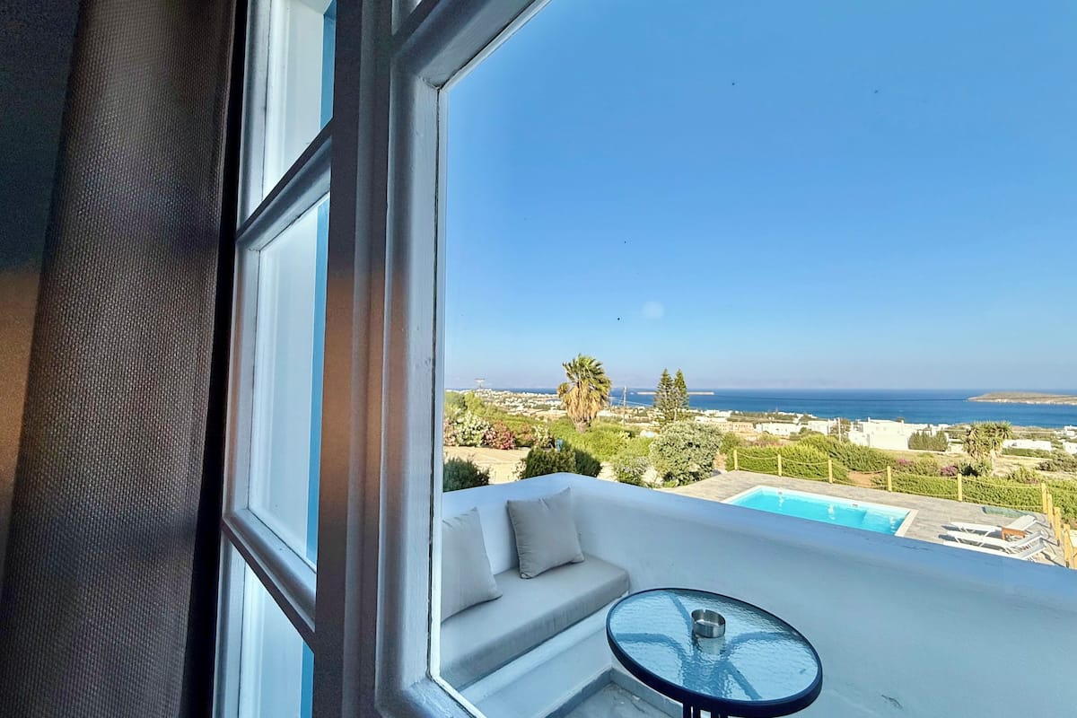 A balcony with a glass table and a sofa, overlooking a pool and a coastal landscape.