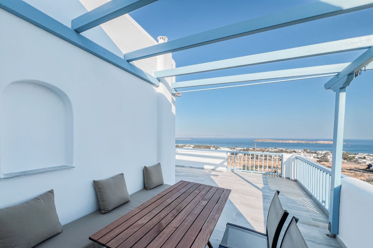 A terrace with a wooden table and chairs, a white wall with a window, and a view of the sea and distant land.