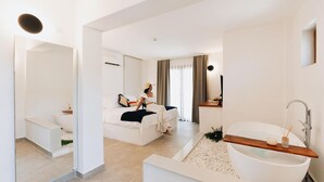 Ett modernt hotellrum med en stor säng, en sittgrupp, ett badrum med badkar och en spegel.