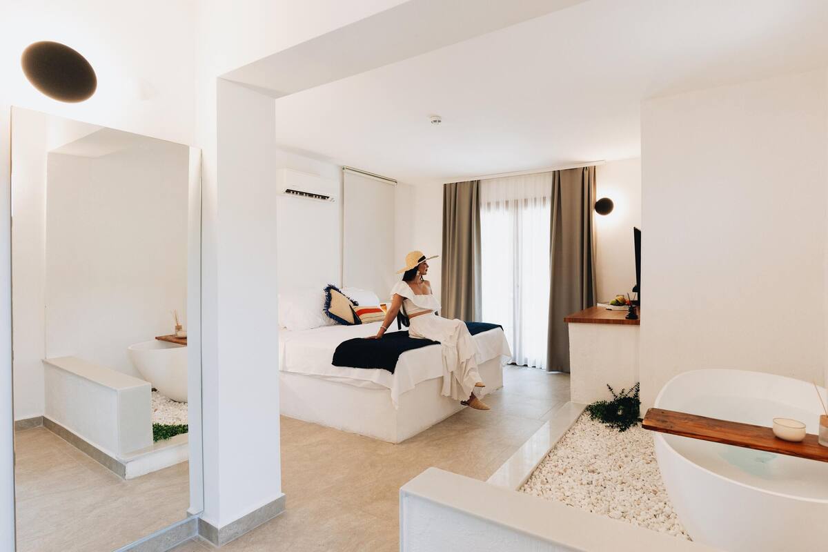 Una habitación de hotel moderna con una cama grande, una zona de estar, un baño con bañera y un espejo.