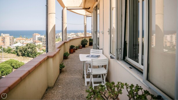 En balkon med et bord og stole, krukker med planter og udsigt til havet og bygninger.