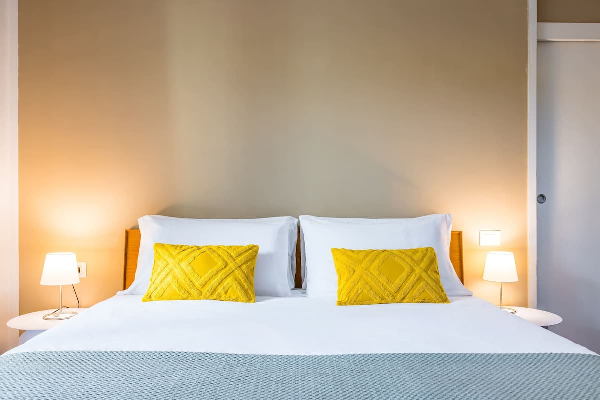 A hotel room with a bed, two yellow pillows, two bedside lamps, and a white nightstand.