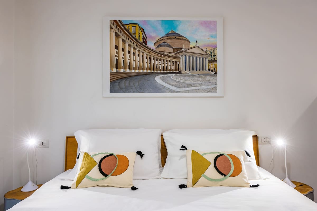 A hotel room with a bed, two bedside lamps, and a painting of a cityscape on the wall.