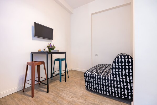 A small, modern room with a desk, two stools, a black and white patterned sofa, and a flat-screen TV mounted on the wall. - Direlo Guest House (Naples)