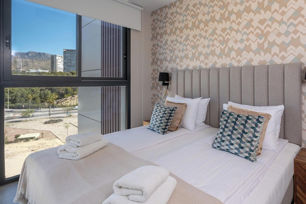 A modern hotel room with a large bed, two side tables, a view of the outdoors, and a patterned wall.